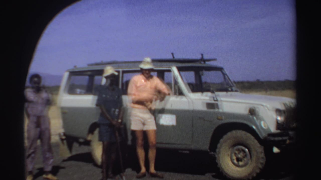In 1969 Sudan, three men stand near a pale green and white Toyota Land Cruiser under a bright sky.