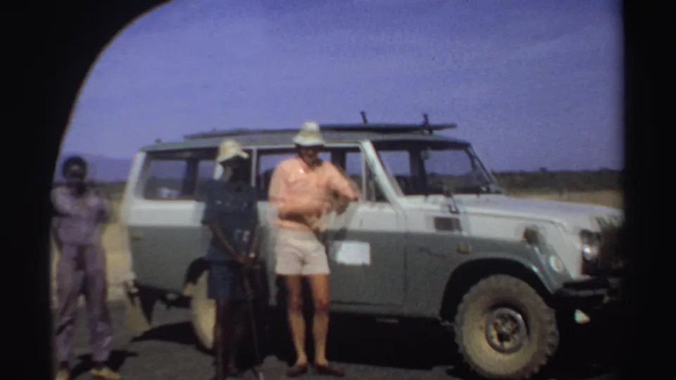 In 1969 Sudan, three men stand near a pale green and white Toyota Land Cruiser under a bright sky.