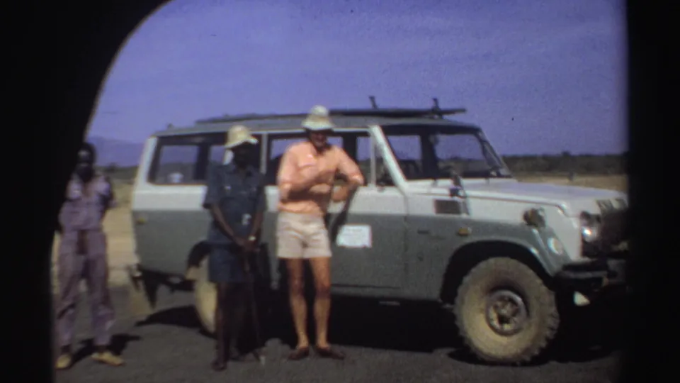 A traveler in a pink shirt and shorts stands beside two men and a Toyota Land Cruiser in Sudan, 1969.
