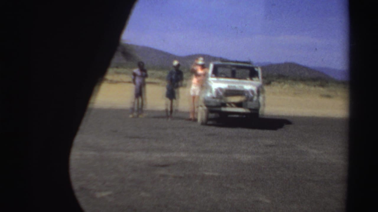 Three people stand beside a white vehicle on an open road with hills behind them.