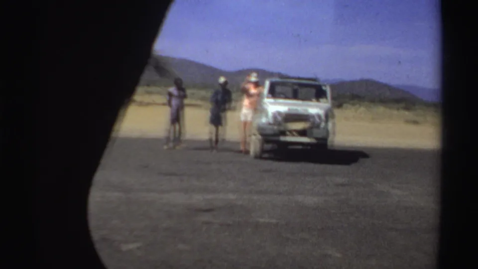 In 1969 Sudan footage, three people stand near a white vehicle on a sunlit road in a sandy, mountainous area.