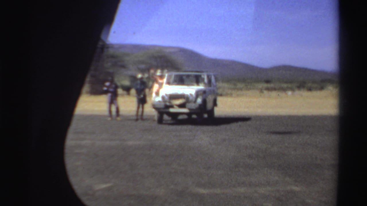 Three people remain near a white vehicle in a dry Sudanese landscape, seen through a soft circular 8mm frame.