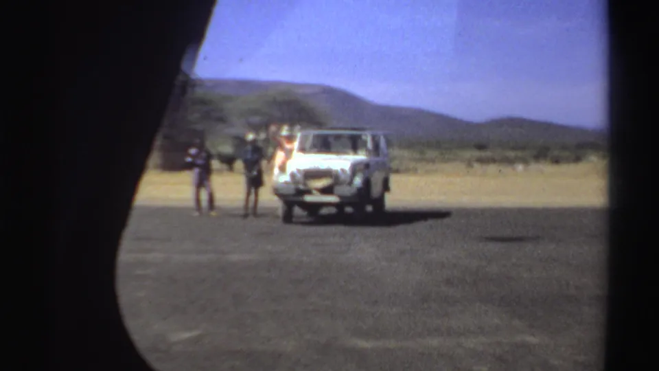 Three people remain near a white vehicle in a dry Sudanese landscape, seen through a soft circular 8mm frame.
