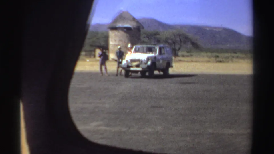 The roadside group and white vehicle appear near a round thatched structure, with hills and sparse trees in the background.