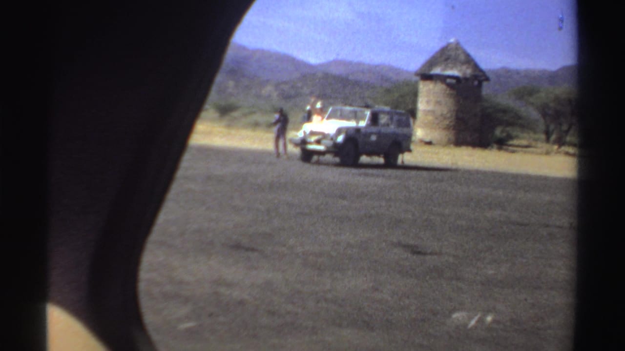 White off-road vehicle parked near a thatched round structure on dusty ground.
