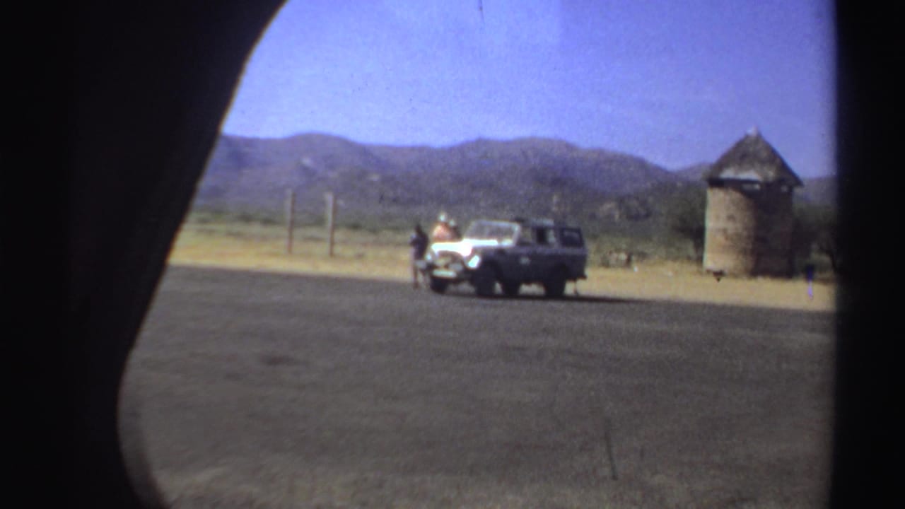White vehicle on open ground near a thatched structure and distant hills.