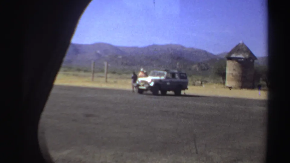 A white off-road vehicle appears parked in a dry Sudan landscape near a round thatched hut, framed from inside another vehicle.