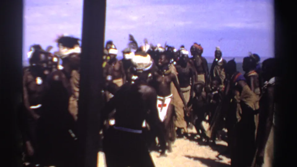 1969 Sudan coastal scene with people in traditional attire dancing or gathering on a beach, partly obscured by dark foreground posts.