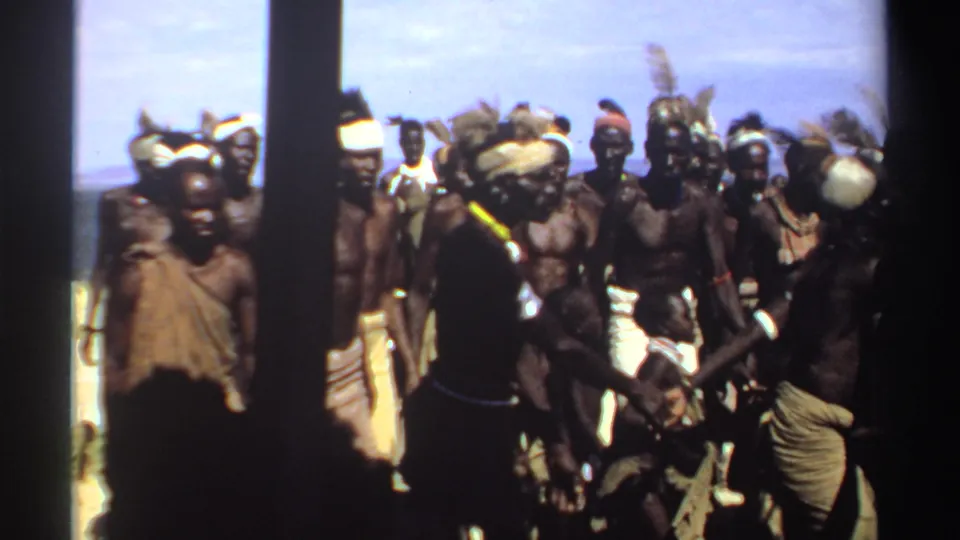 1969 Sudan 8mm frame showing a dense group of men in headbands, jewelry, and wraps during an outdoor dance.