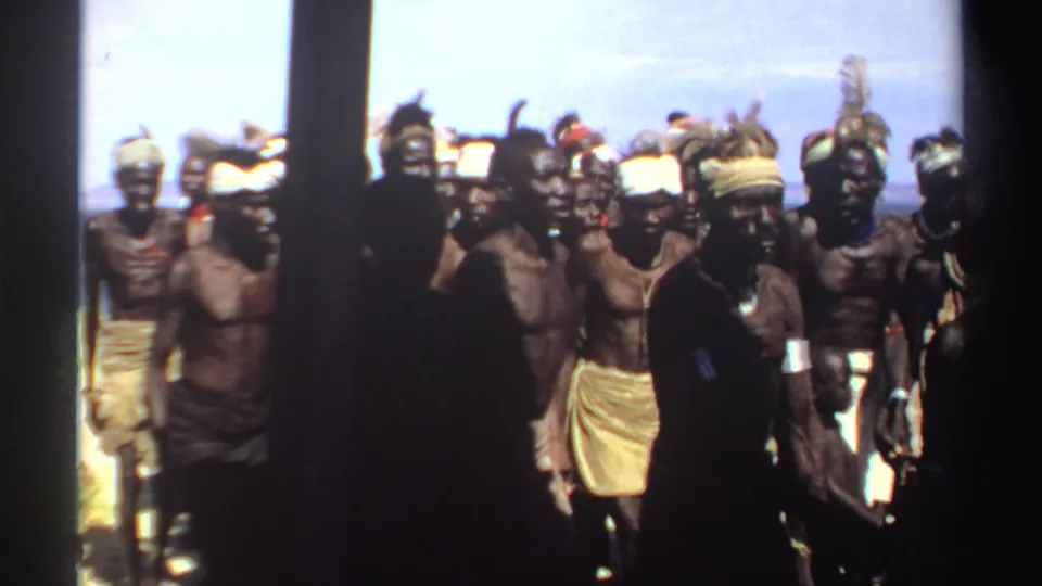 1969 Sudan home-movie frame showing men in wraps, headbands, and feathered ornaments during an outdoor group dance.