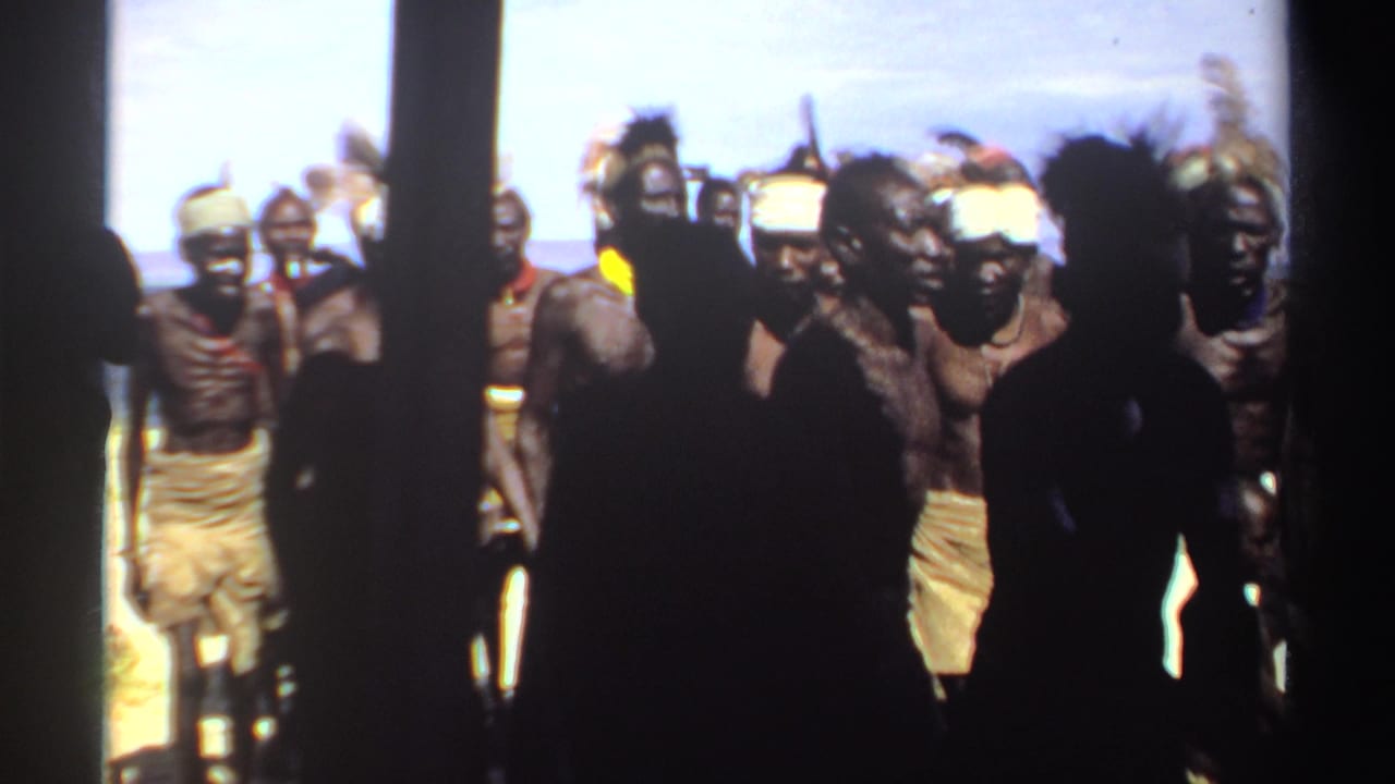 Men in feather headdresses walk outdoors while foreground spectators appear in silhouette.