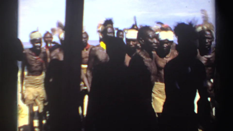 In apparent 1969 Sudan, men wearing feather headdresses and cloth wraps move together outdoors as dark foreground figures obscure part of the view.