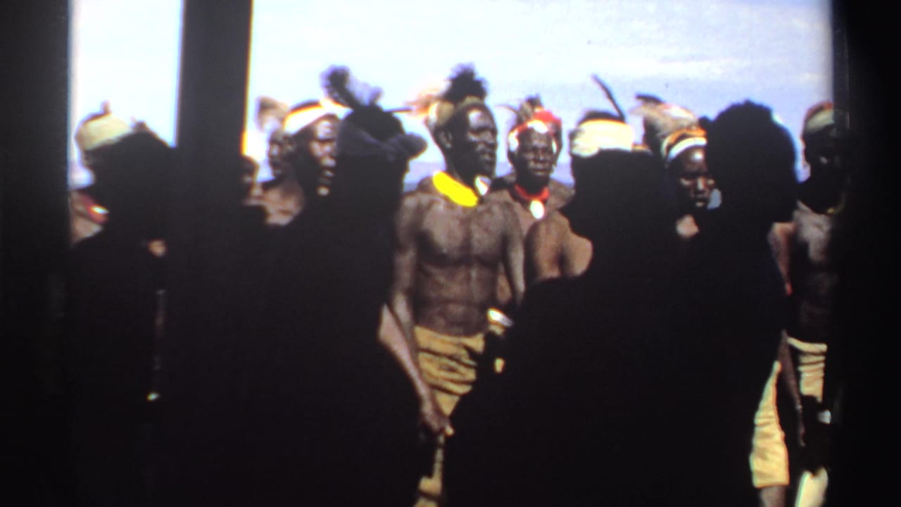 Men in feathered headwear and cloth wraps pass through bright daylight, with faces visible and nearby spectators blocking part of the frame.