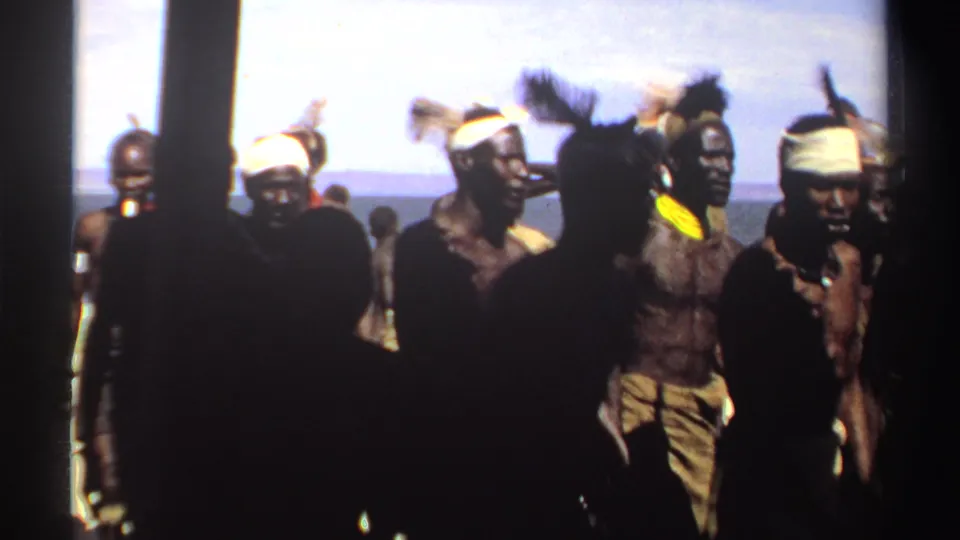 The 8mm frame shows men in ceremonial dress near a distant horizon, partly hidden by dark spectators close to the camera.
