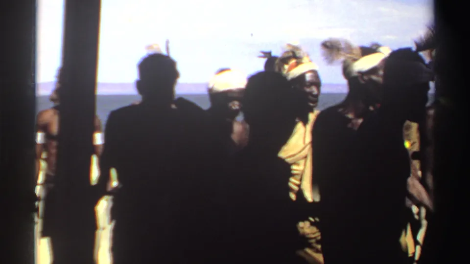 Near the end of the clip, dark foreground figures block much of the view while men in feathered headwear remain visible in the background.