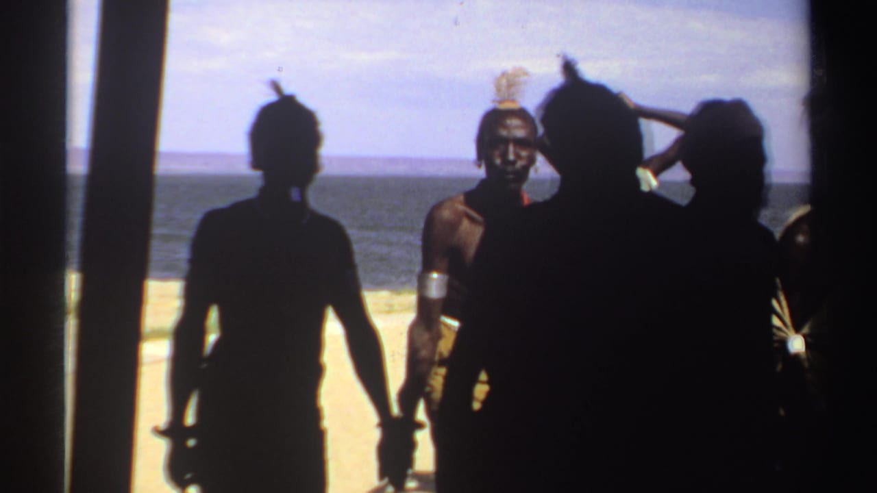 Backlit 1969 Sudan shoreline frame with silhouetted villagers, feathered headwear, arm bands, sandy ground, and open water beyond.