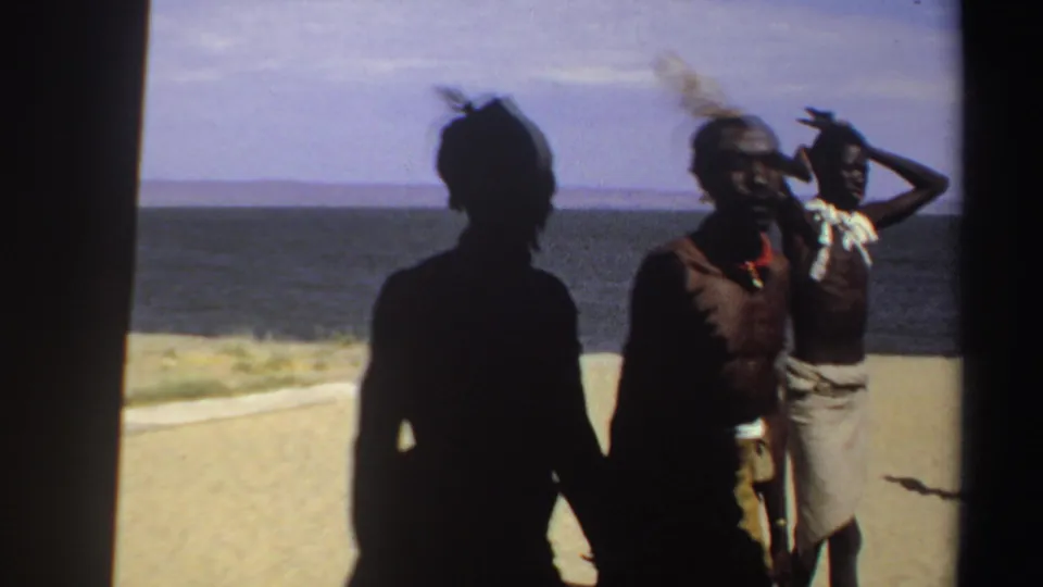 1969 Sudan 8mm frame with two villagers by the shoreline, one looking toward the camera and another raising a hand near his head.