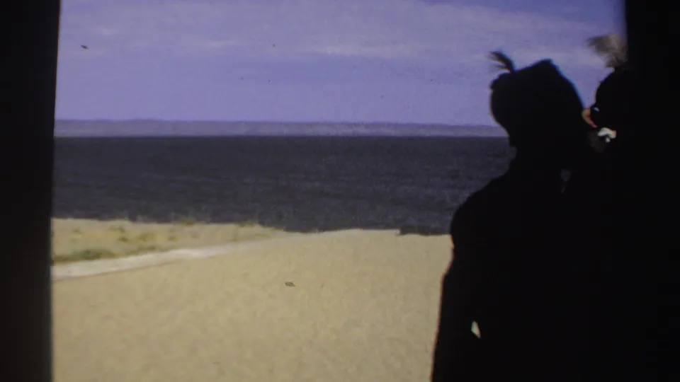 1969 Sudan shoreline frame with silhouetted villagers at right, sandy foreground, calm water, and a distant landform across the horizon.