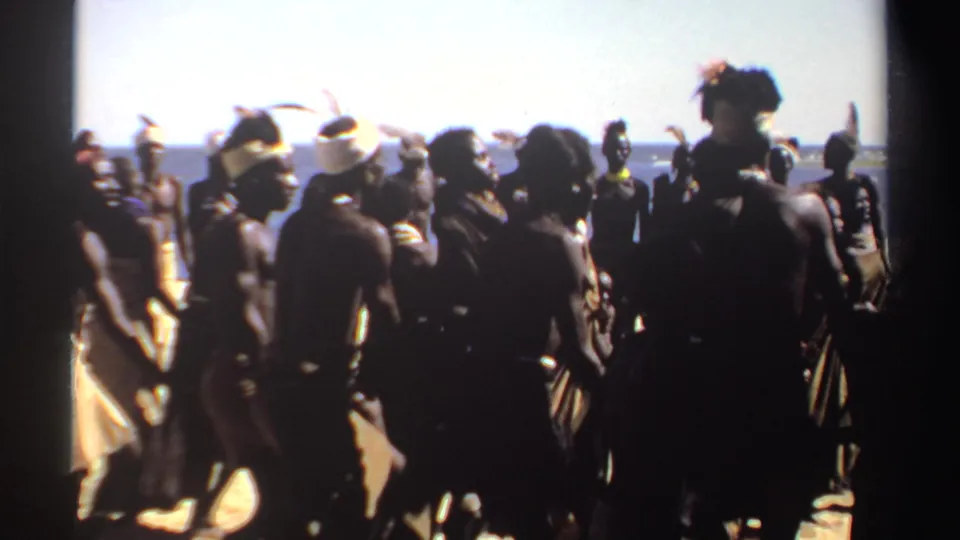 1969 Sudan 8mm frame showing a coastal group dance with silhouetted figures, feathered headbands, and wrapped garments.
