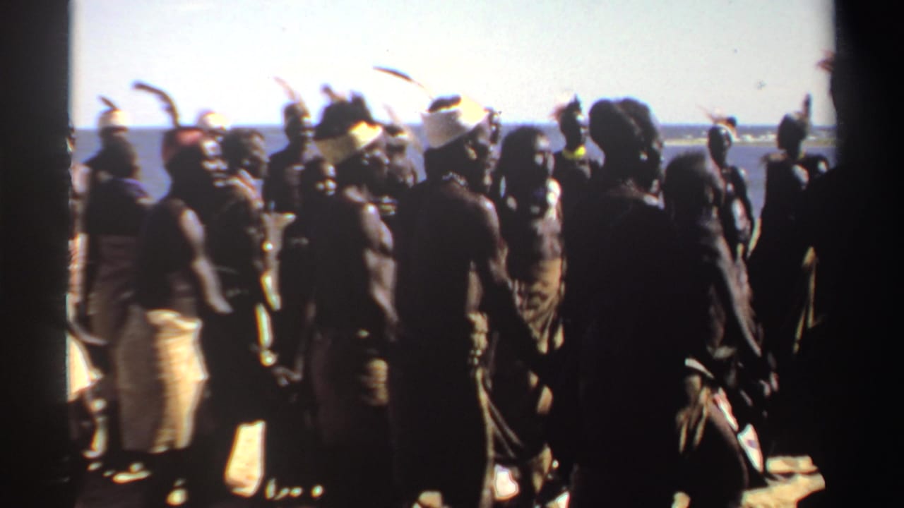 1969 Sudan home-movie frame of dancers in head coverings and feathered adornments moving along a bright coastal shoreline.