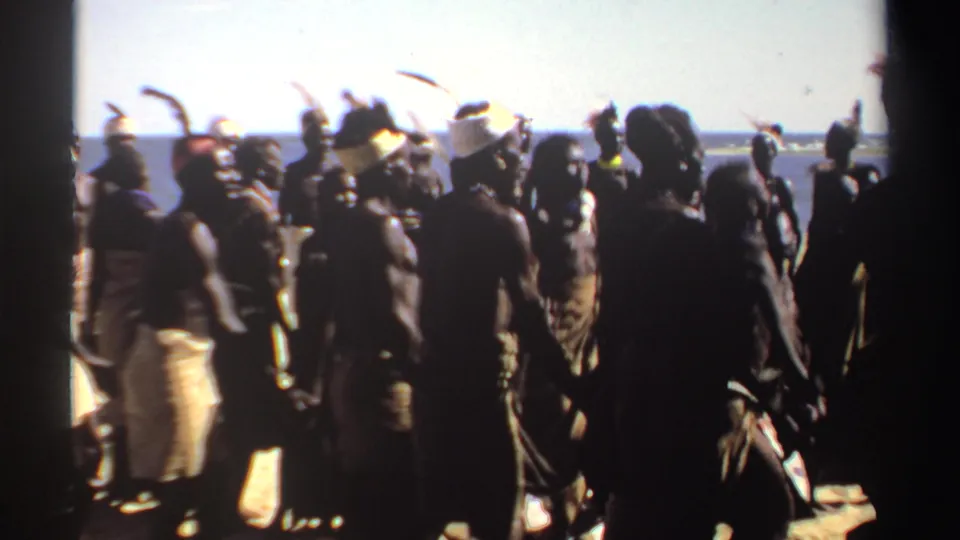 1969 Sudan home-movie frame of dancers in head coverings and feathered adornments moving along a bright coastal shoreline.