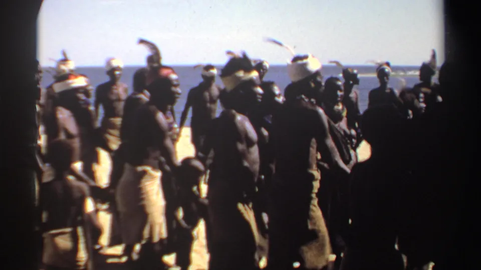 1969 Sudan 8mm frame with a handheld view of dancers in wrapped cloth and head adornments near the ocean.