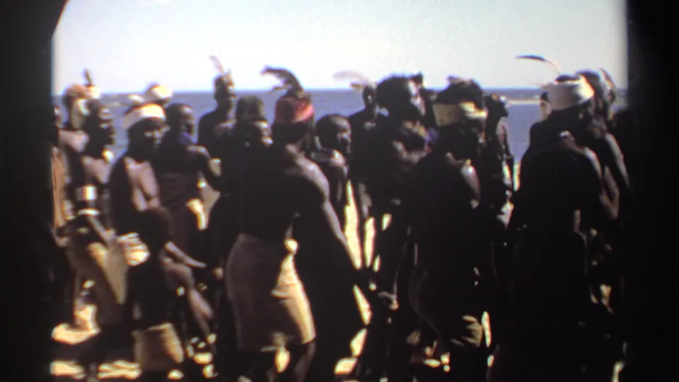 1969 Sudan home-movie frame showing a dense group of dancers silhouetted against a bright coastal background.