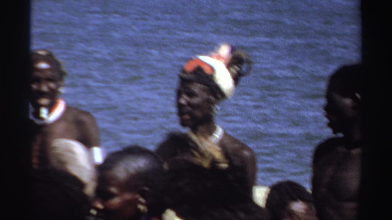 1969 Sudan 8mm frame showing men with feathered headgear, necklaces, light cloth wraps, and white pigment standing near blue water.