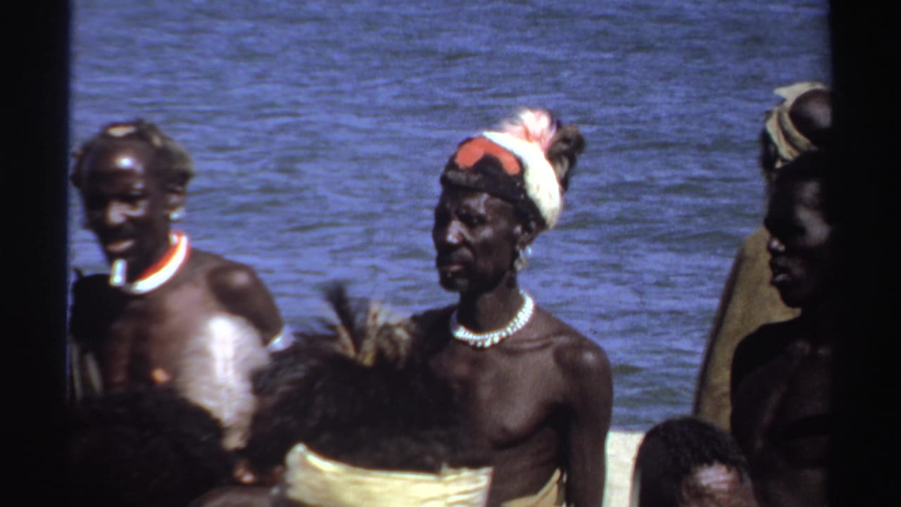 Several men stand near water, including one with a feathered headpiece and necklace.