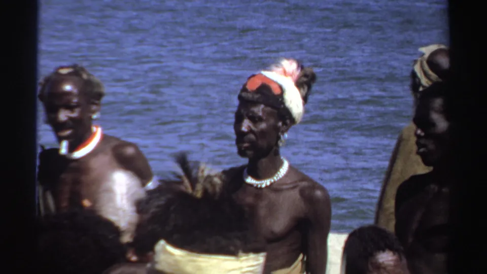 1969 Sudan frame with men near blue water, one wearing a feathered headpiece and necklace, with white pigment visible on another man's torso.