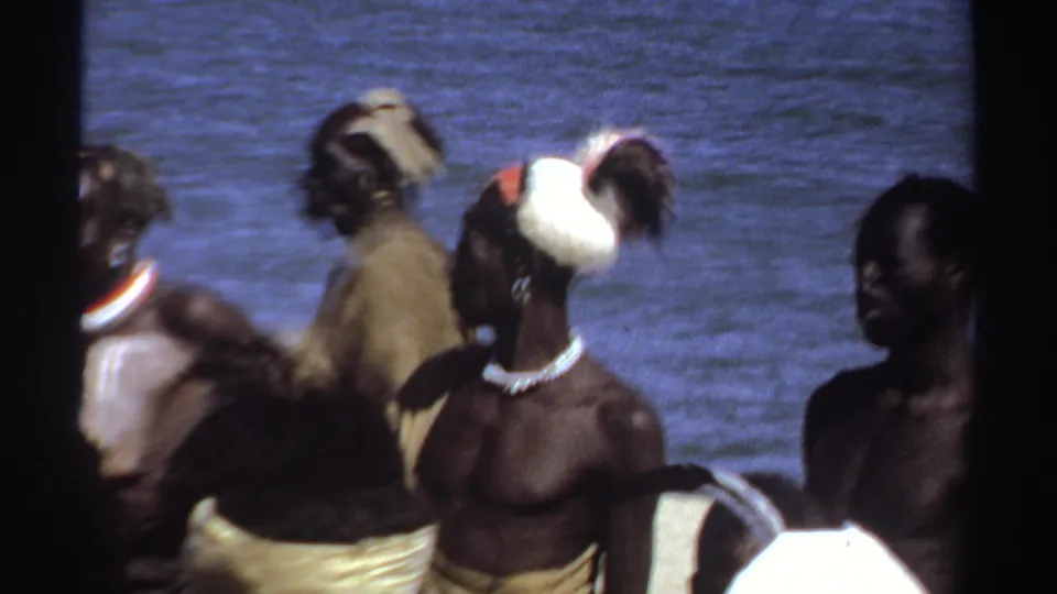 1969 Sudan 8mm frame showing men shifting position near water, including a feathered headpiece, necklaces, cloth wraps, and white pigment markings.