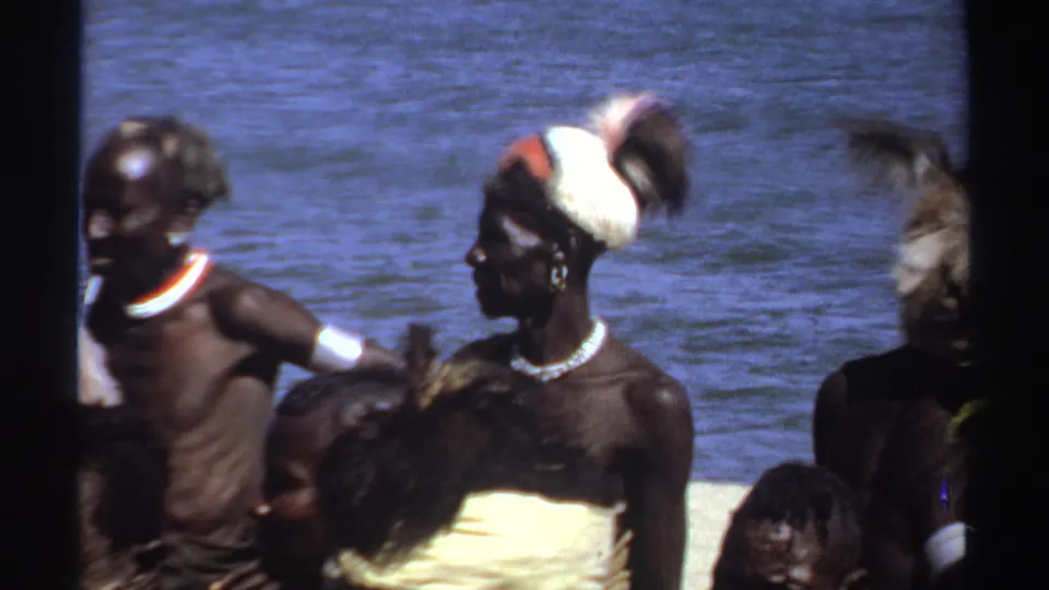 1969 Sudan frame of men at a waterfront, with feathered headgear, white pigment, necklaces, and light wraps visible in a soft 8mm image.