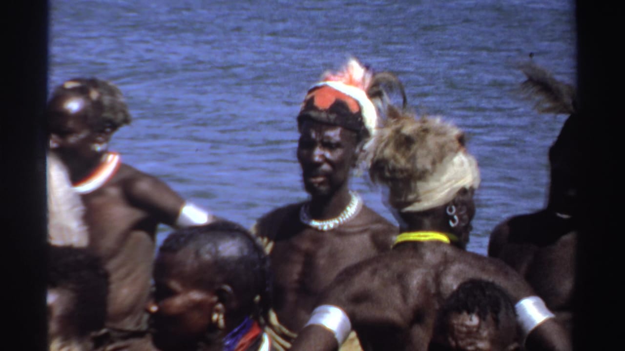 1969 Sudan 8mm frame showing adults in traditional adornment gathered by water, partly blocked by dark foreground edges.