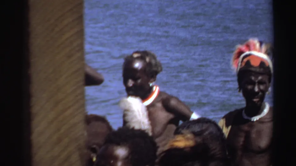 1969 Sudan 8mm frame showing adults in traditional adornment near blue water, with the camera partly blocked at left.