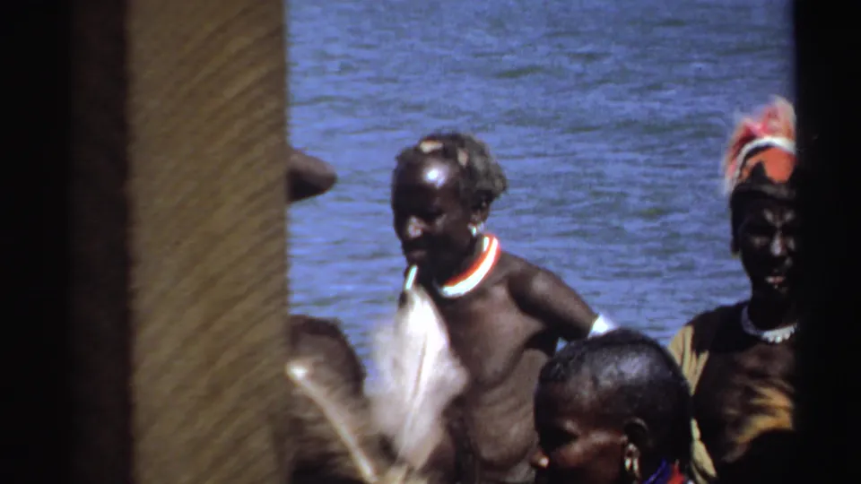 1969 Sudan frame with adults in feathered headwear and necklaces beside rippling water, framed by dark and tan foreground edges.