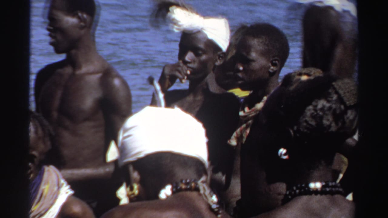 1969 Sudan home-movie frame showing people standing close together near water, with headwraps, beads, and sunlit faces visible.