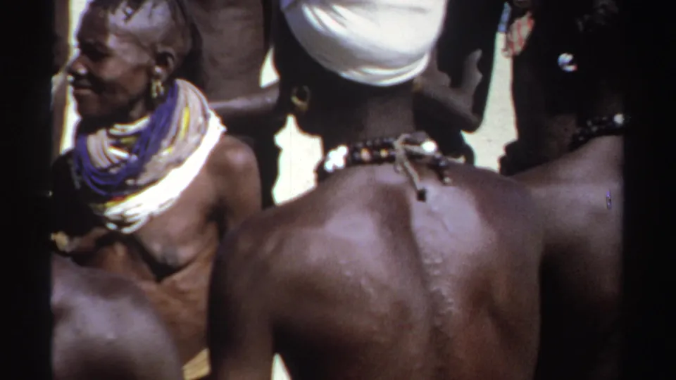 1969 Sudan frame showing an older adult in profile within a close outdoor gathering, with headwraps and beaded necklaces nearby.