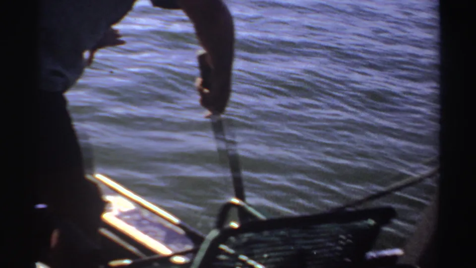 1969 Sudan 8mm frame of a man leaning over a small boat side, lowering or holding a pole or line beside the water.