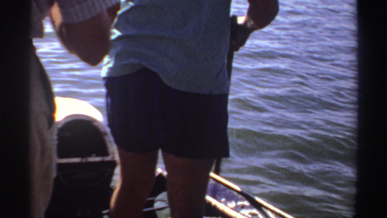 1969 Sudan home-movie frame of an adult in a light shirt and dark shorts holding a fishing rod on a small boat.