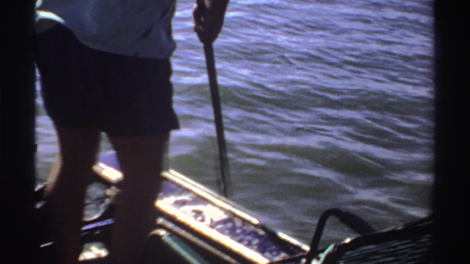 1969 Sudan 8mm frame with a close handheld view of an adult holding a fishing rod beside sunlit water.