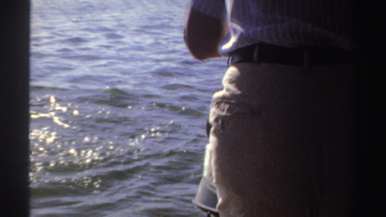 1969 Sudan home-movie frame with an adult in a striped shirt near the side of a boat, facing sunlit water.