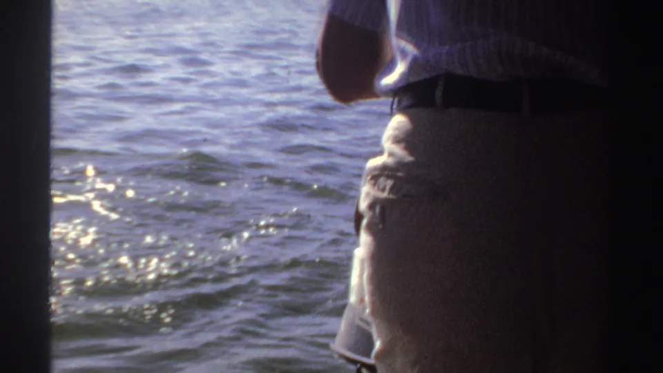 1969 Sudan home-movie frame with an adult in a striped shirt near the side of a boat, facing sunlit water.