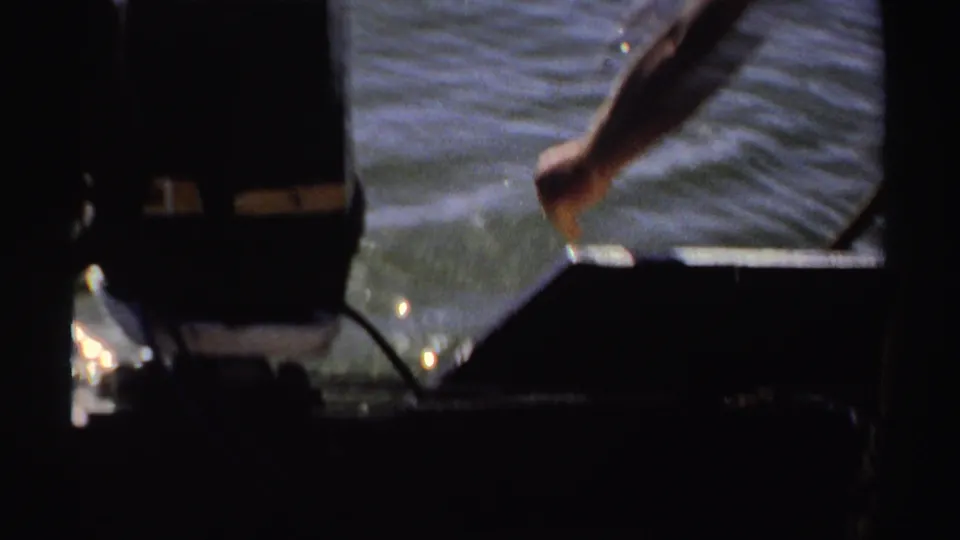 1969 Sudan 8mm frame showing an adult arm extended over a boat edge toward rippling lake water.