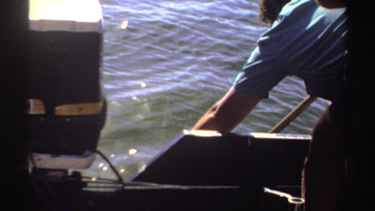 1969 Sudan 8mm frame showing an adult leaning over a boat edge with one arm extended toward sunlit water near a dark outboard motor.