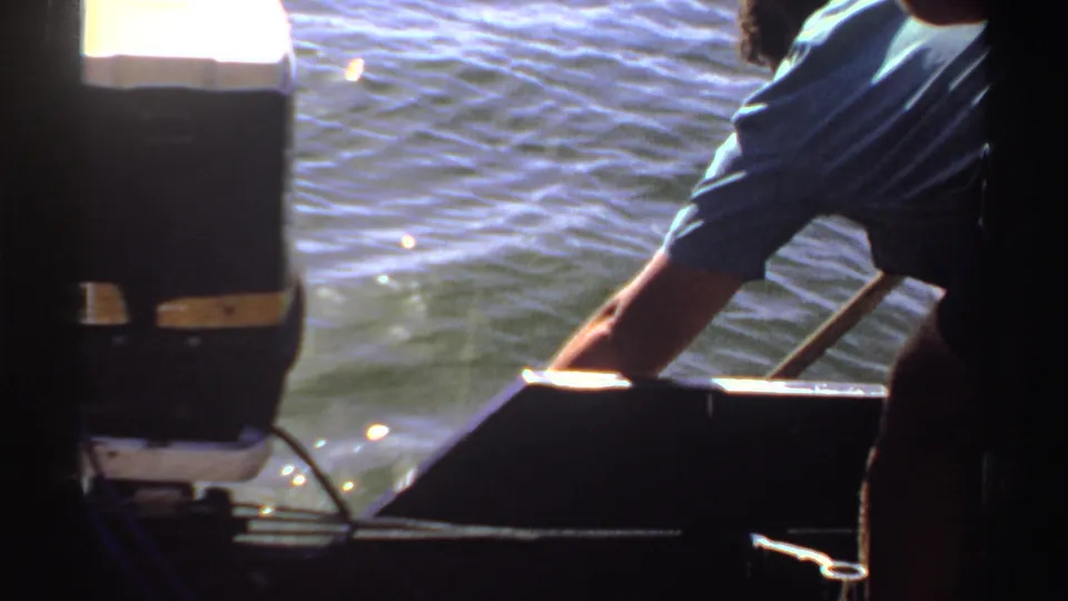 1969 Sudan 8mm frame showing an adult leaning over a boat edge with one arm extended toward sunlit water near a dark outboard motor.