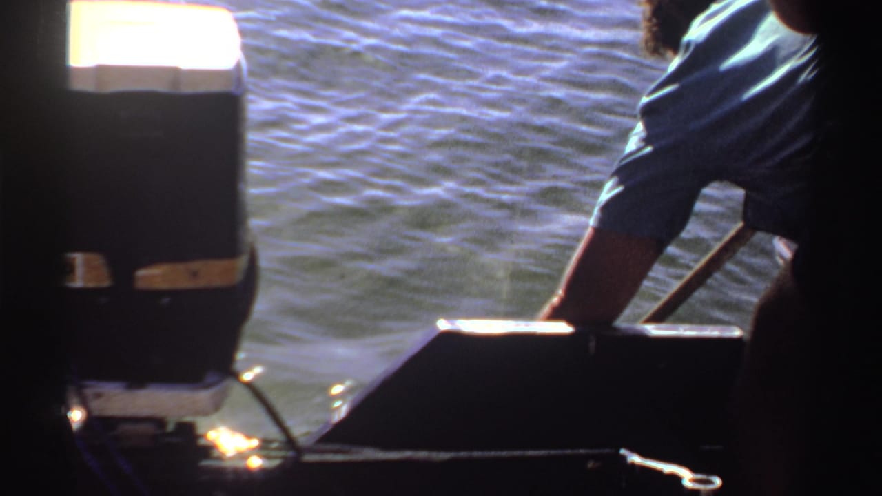 1969 Sudan 8mm frame with a partial adult in a blue shirt leaning from the boat as a pole angles toward the water.
