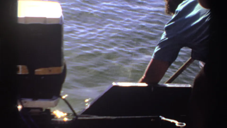 1969 Sudan 8mm frame with a partial adult in a blue shirt leaning from the boat as a pole angles toward the water.