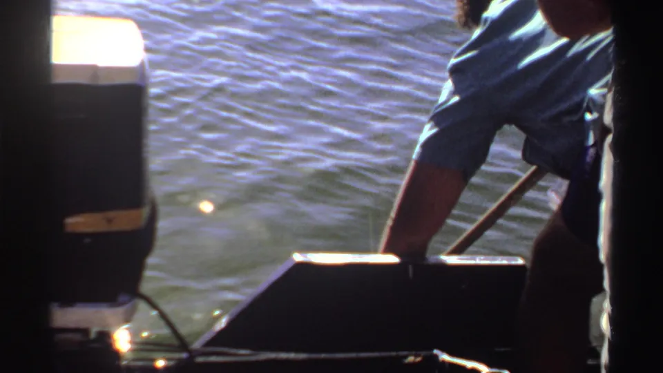 1969 Sudan 8mm frame showing a blue-sleeved adult reaching down over a dark boat side as sun flashes on the water surface.