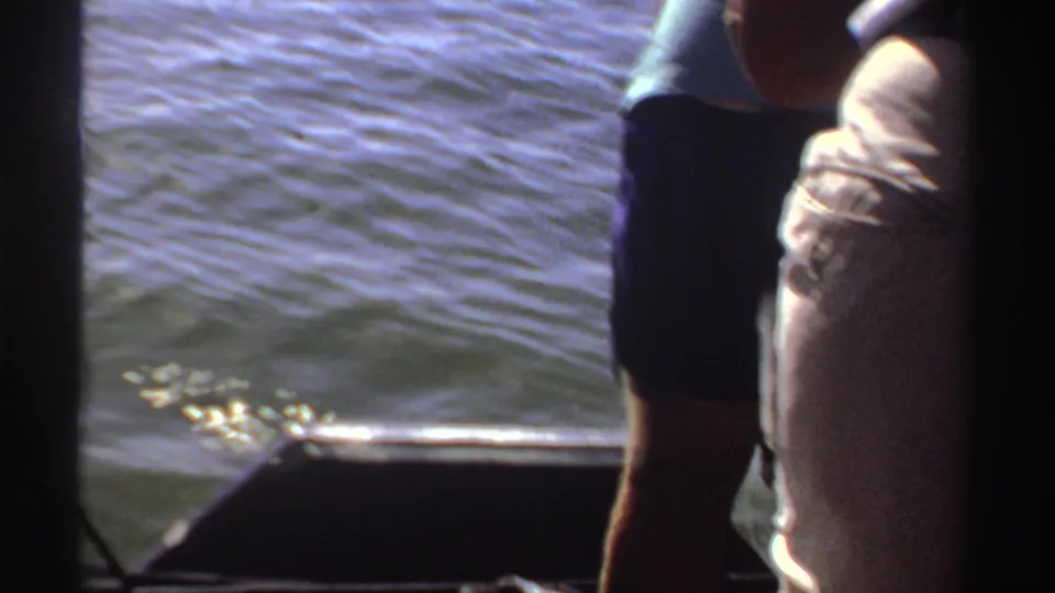 1969 Sudan 8mm frame showing partial standing adults near the side of a small boat, with rippled water and motor hardware nearby.