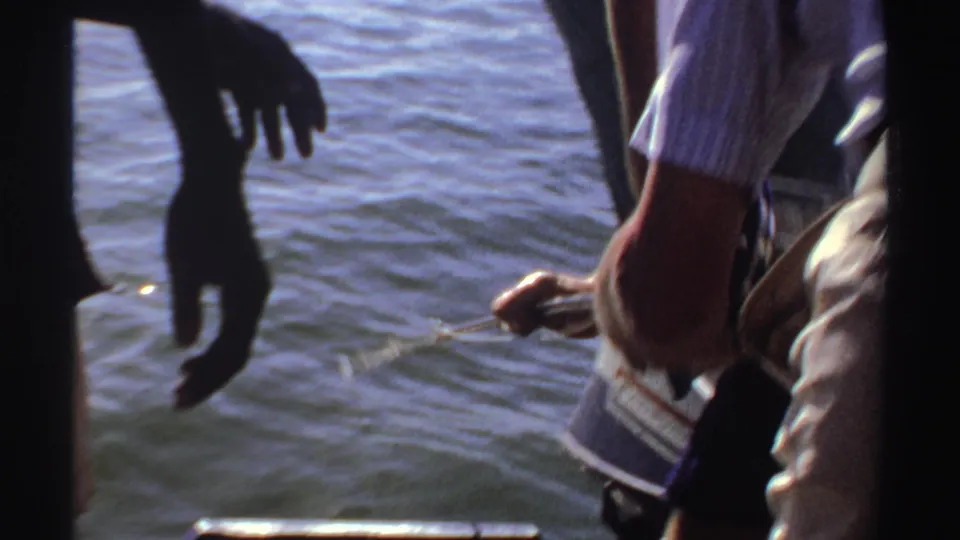 1969 Sudan home-movie frame showing a man leaning down near the boat edge as another adult stands beside sunlit water.